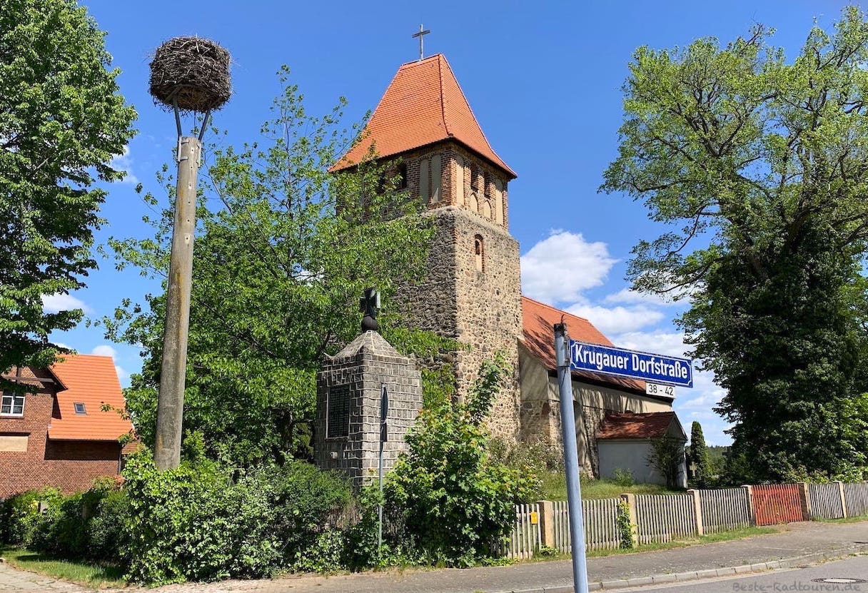 Foto vom Radweg her: Kirche von Krugau, Storchennest und Kriegsdenkmal